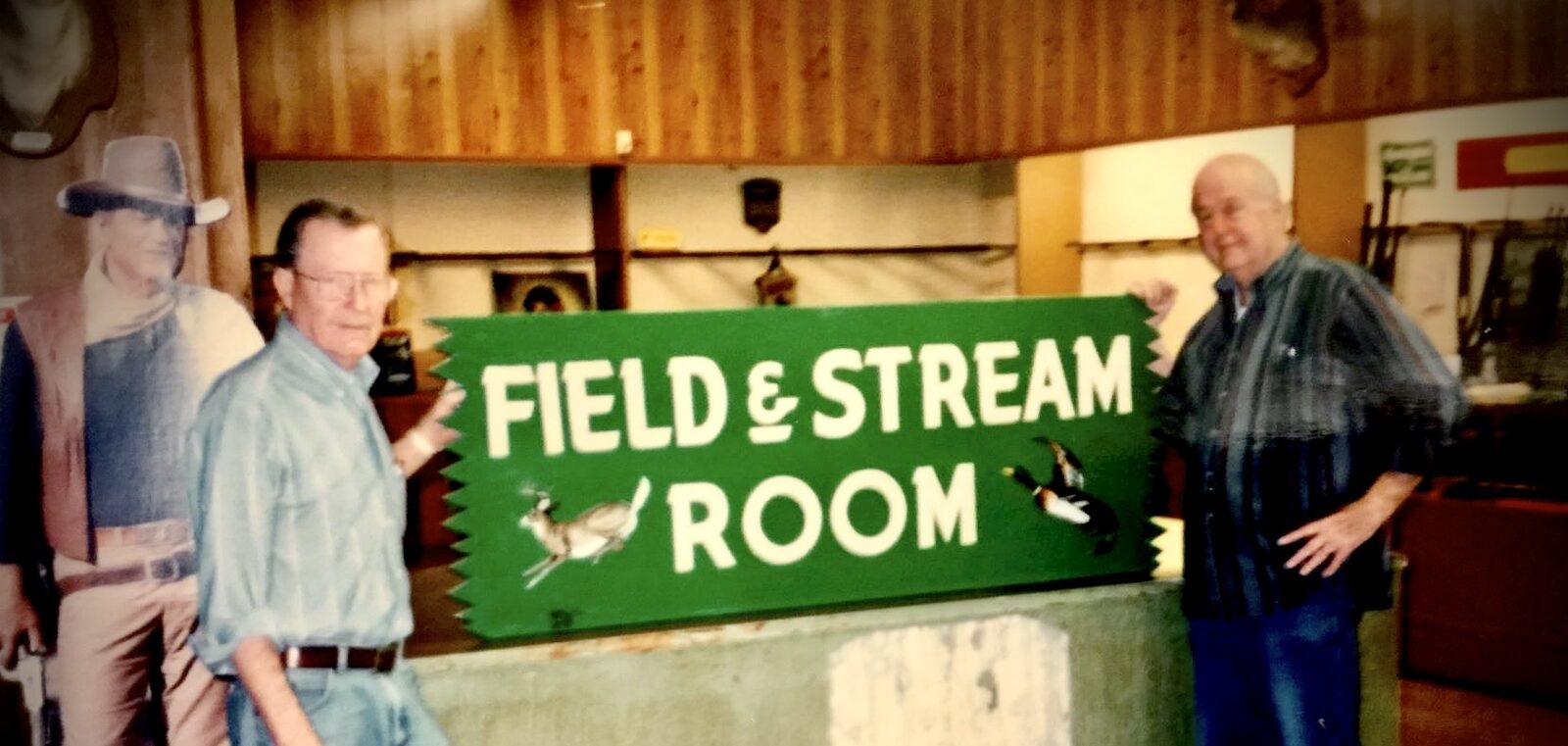 Leo Bradshaw and another man holding the green Field and Stream Room sign inside the shop.